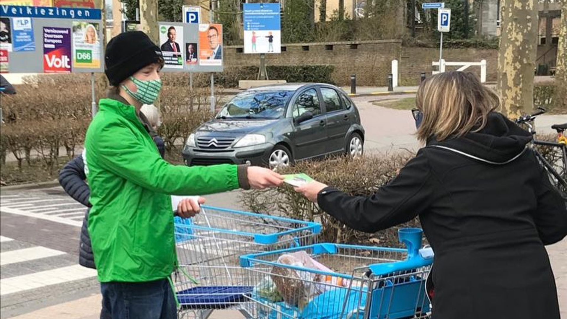 Persoon in groen jasje deelt folder uit aan persoon met volle winkelwagen